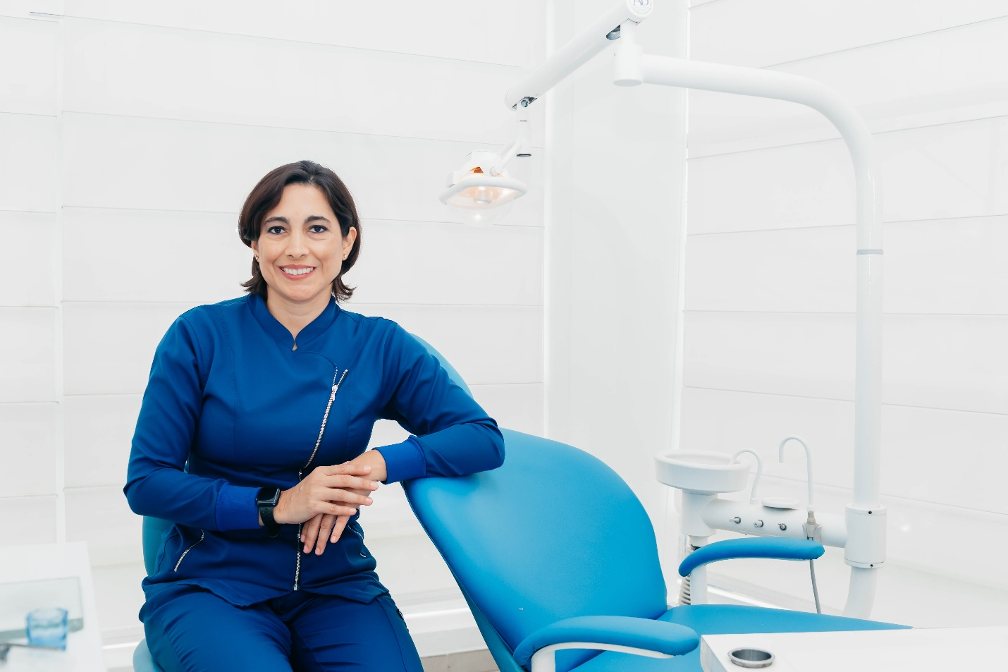 A dentist wearing blue smiling at the camera while sitting beside the dentist’s chair.