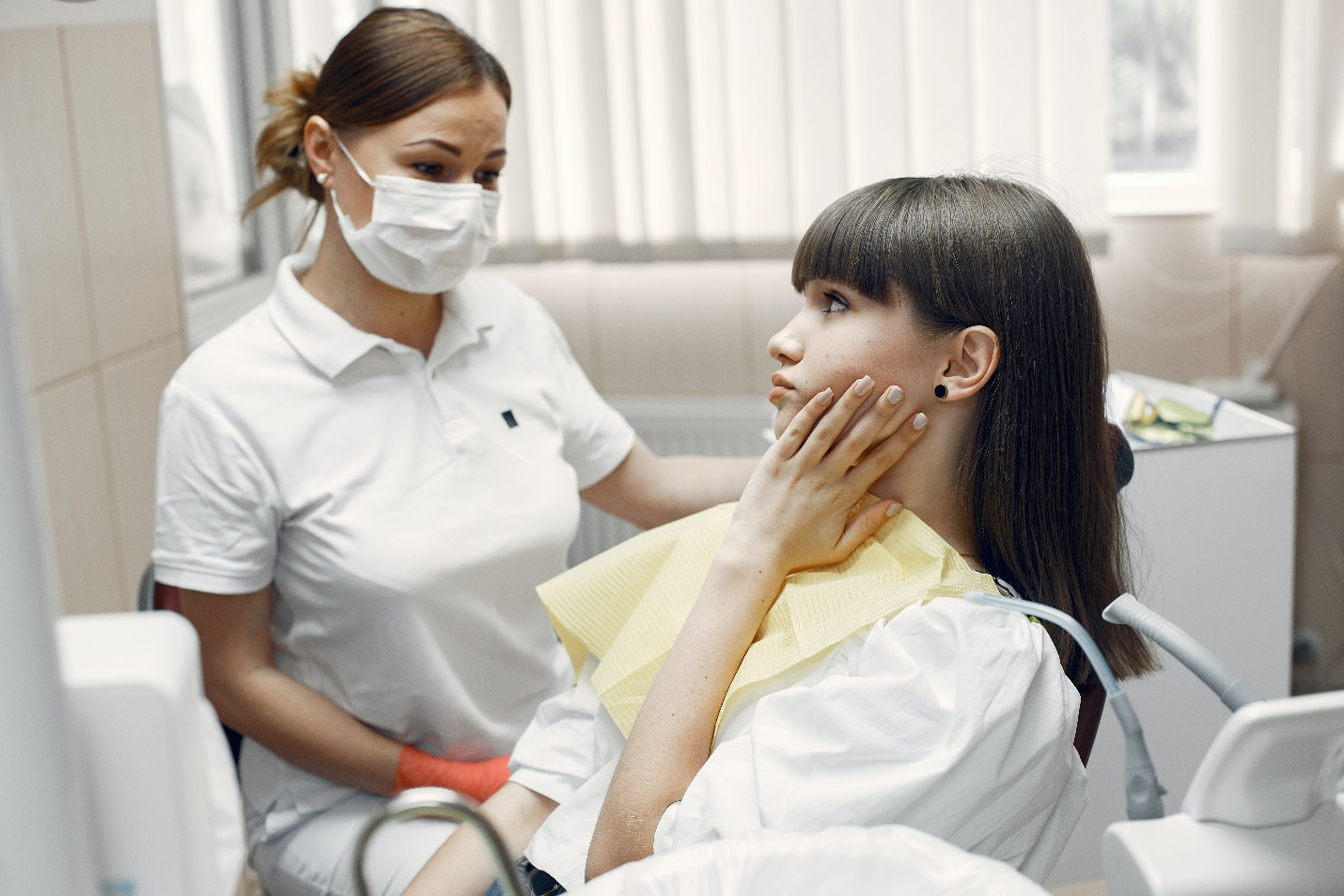 A woman at the dentist’s holding her left cheek while someone attends to her.